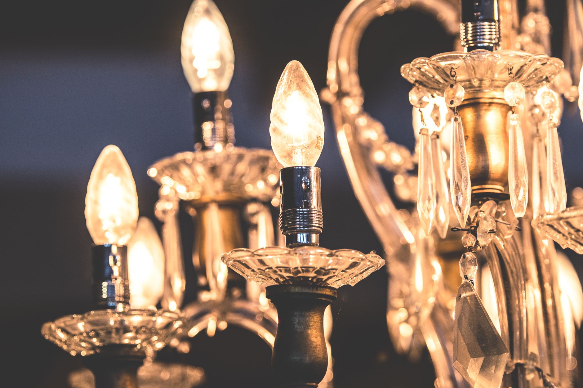 Gros plan d’un lustre en cristal avec ampoules allumées, bobèches en verre et pendeloques taillées, sur fond sombre.