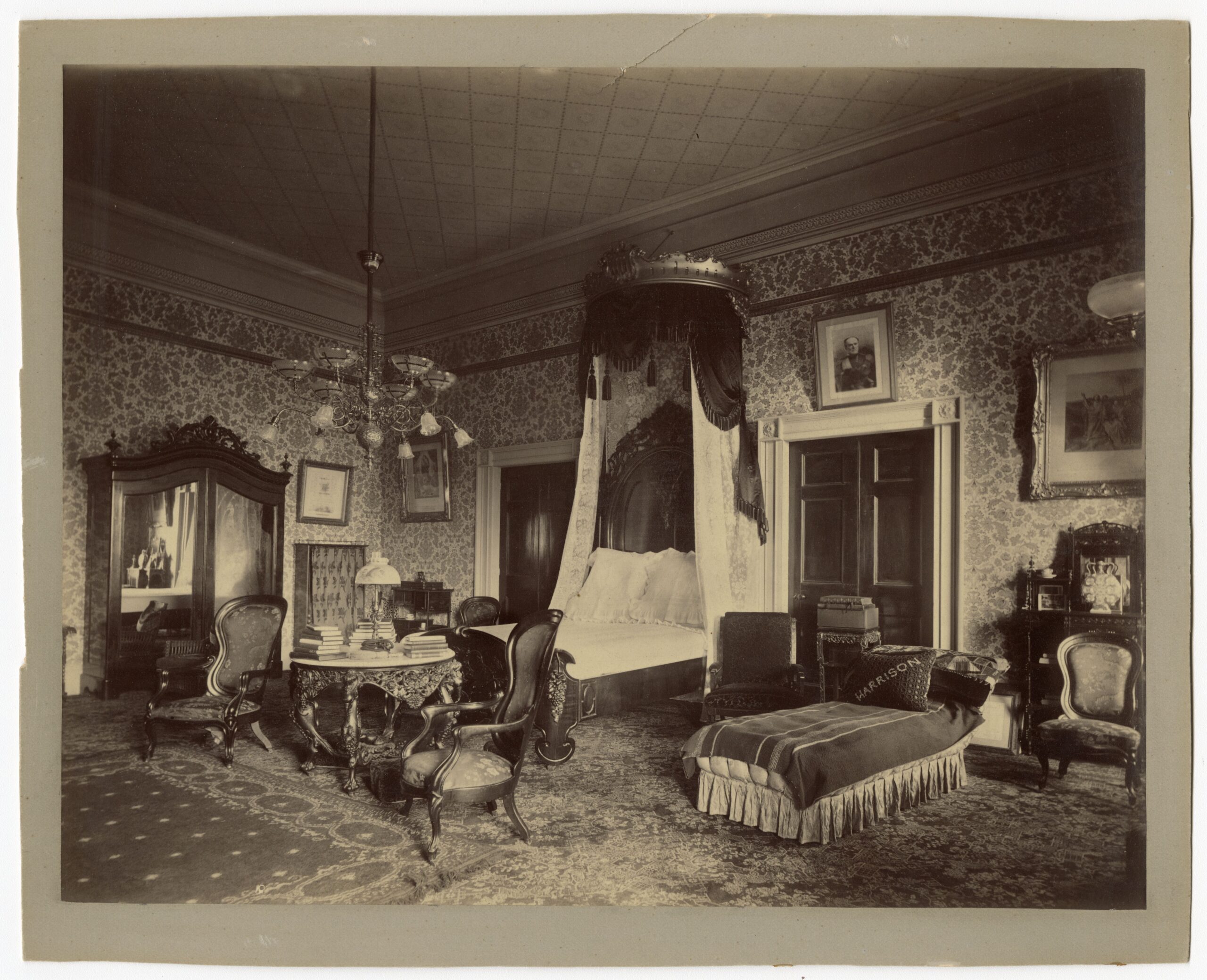 Photographie ancienne d’une chambre avec lit à baldaquin, papier peint floral, armoire à glace, fauteuils, table ronde et lustre.