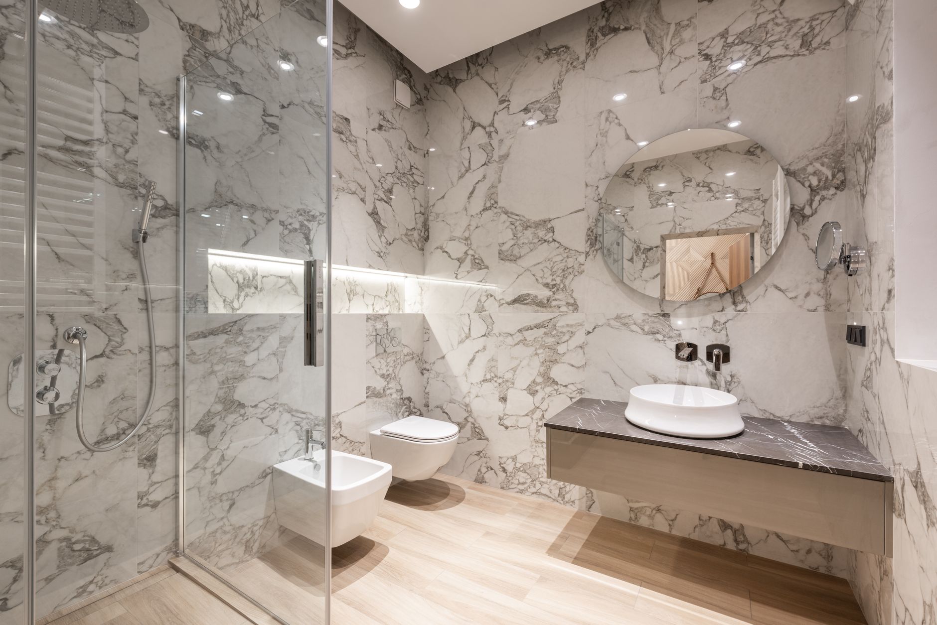 Salle de bain moderne en carrelage effet marbre avec douche vitrée, WC et bidet suspendus, miroir rond et vasque sur plan suspendu.