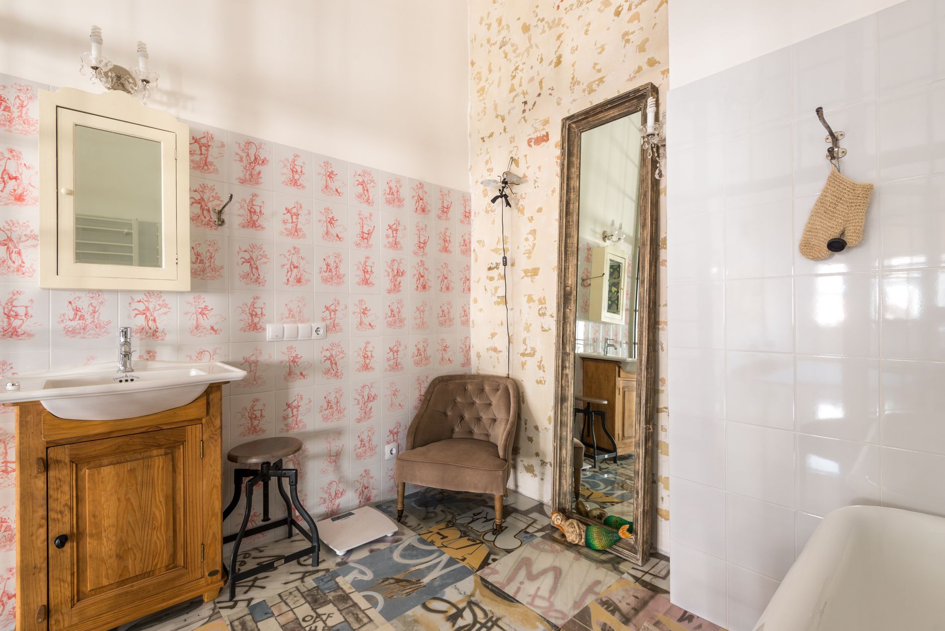 Salle de bains au carrelage mural à motifs rouges, avec meuble vasque en bois, grand miroir, fauteuil capitonné et sol en carreaux colorés.